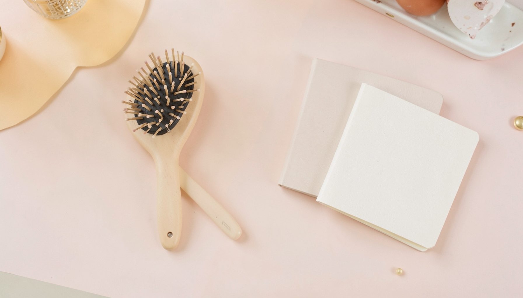 Flat lay of fashion accessories, a hairbrush, and a small notebook on a pastel desk, warm and feminine mood