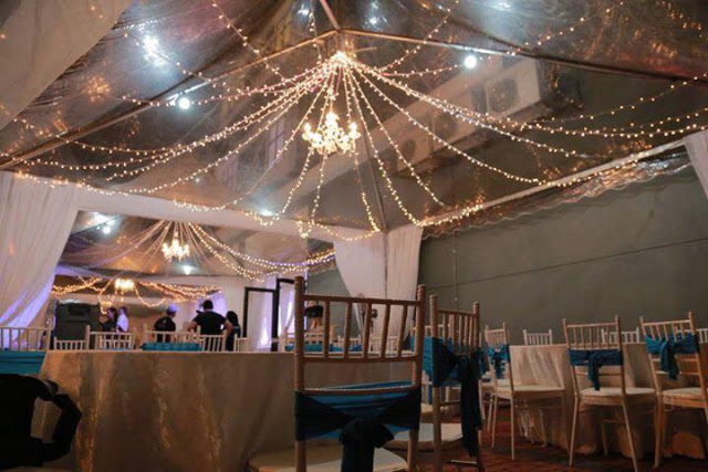 Event tent interior with string lights and chandeliers hanging from silver drapery, chairs with blue sashes, warm festive mood