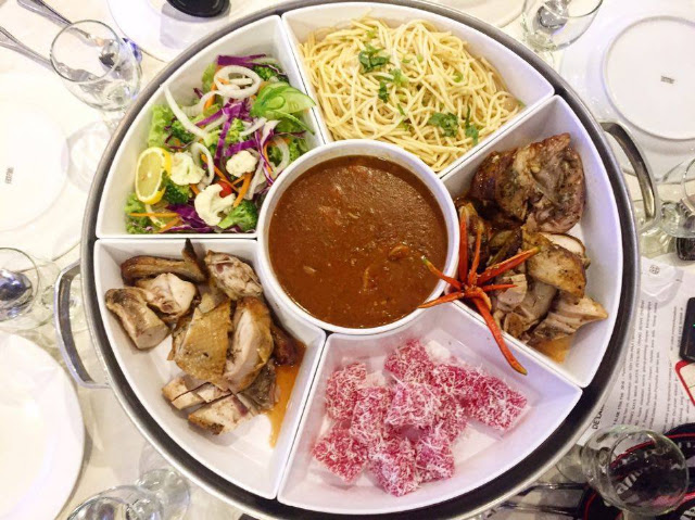 Round platter divided into compartments with pasta, salad, meat, sauce, and dessert bites, on a table with glasses, warm